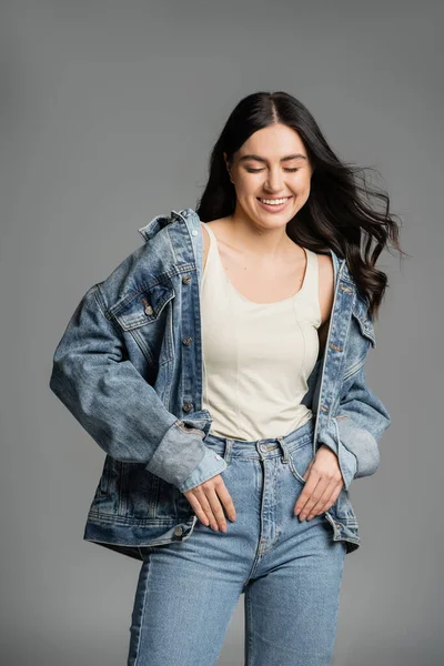 Happy young woman with gorgeous brunette hair posing in stylish blue jeans and denim jacket while smiling with closed eyes on grey background — Stock Photo