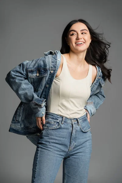 Happy young woman with gorgeous brunette hair posing with hands on waist and standing in stylish blue jeans and denim jacket while looking away on grey background — Stock Photo