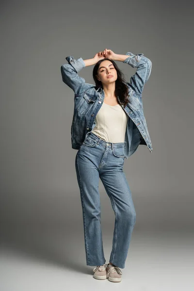 Повна довжина шикарної жінки з брюнеткою волосся позує руками над головою і закритими очима, стоячи в стильних блакитних джинсах і джинсовій куртці на сірому фоні — стокове фото