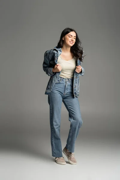 Full length of enchanting woman with brunette hair smiling with closed eyes while posing in stylish blue jeans and denim jacket and standing on grey background — Stock Photo