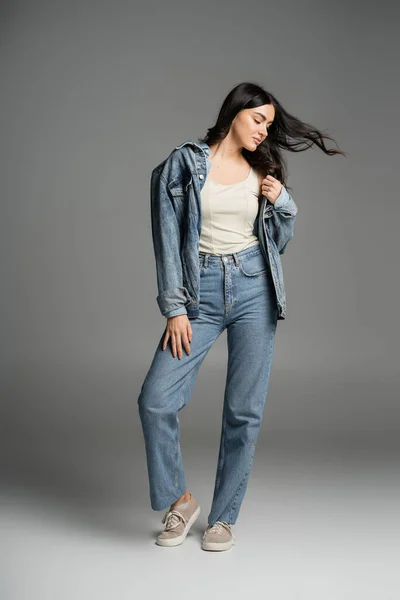 Полная длина очаровательной женщины с длинными брюнетками волосы машут от ветра позируя в стильных синих джинсах и джинсовой куртке, стоя на сером фоне — стоковое фото