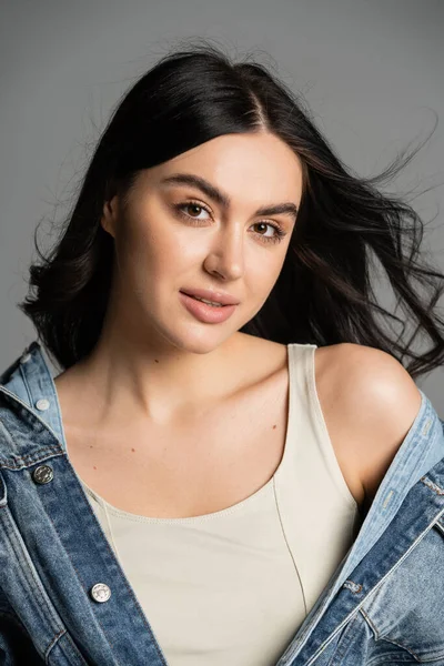 Portrait of charming young woman with brunette hair posing in fashionable and blue denim jacket and looking at camera isolated on grey background — Stock Photo