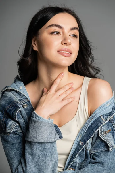 Portrait of charming woman with brunette hair posing in stylish and blue denim jacket and holding hand near neck while looking away isolated on grey background — Stock Photo