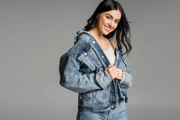 Portrait of cheerful young woman with brunette hair posing in stylish denim jacket and blue jeans while smiling and standing isolated on grey background — Stock Photo