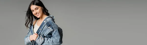 Ritratto di giovane donna allegra con bruna e splendidi capelli in posa in giacca di jeans elegante mentre sorride e in piedi isolato su sfondo grigio, banner — Foto stock