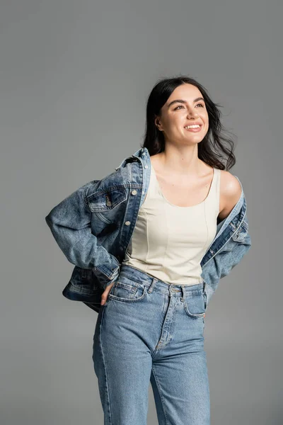 Happy young woman with brunette hair standing in blue jeans and stylish denim jacket while smiling and posing with hand on hip isolated on grey background — Stock Photo