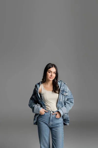 Брюнетка молодая женщина с великолепными волосами стоя с руками в карманах синих джинсов и позируя в стильной джинсовой куртке, глядя на камеру изолированы на сером фоне — стоковое фото