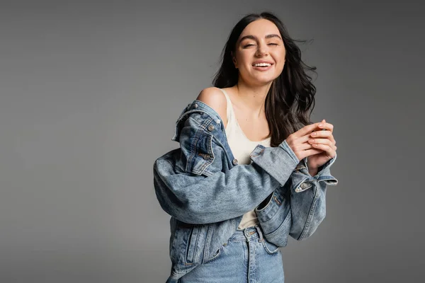 Giovane donna positiva con capelli castani in piedi in jeans blu e giacca di jeans alla moda mentre sorride e posa isolata su sfondo grigio — Foto stock
