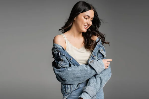 Alluring young woman with brunette hair and natural makeup standing in blue jeans and fashionable denim jacket while smiling and posing isolated on grey background — Stock Photo