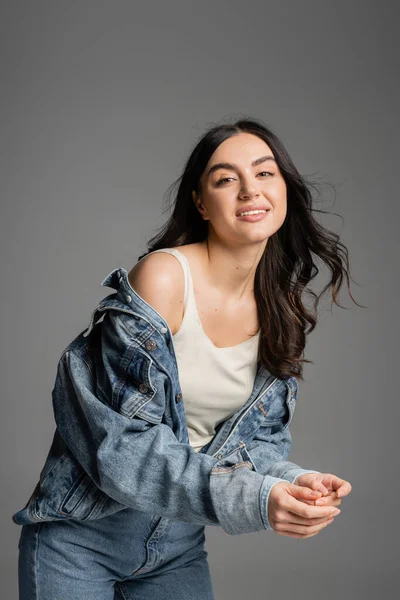 Happy young woman with brunette hair and flawless natural makeup standing in fashionable denim jacket while smiling while posing isolated on grey background — Stock Photo