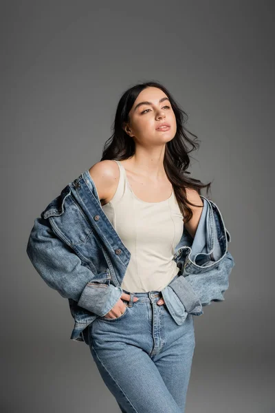 Brunette young woman with shiny hair standing with hands in pockets of blue jeans and posing in stylish denim jacket while looking away isolated on grey background — Stock Photo
