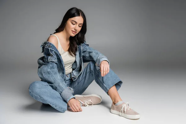 Full length of young charming woman with long brunette hair and flawless natural makeup posing in stylish blue jeans and denim jacket while sitting on grey background — Stock Photo