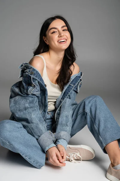 Full length of happy woman with long brunette hair and flawless natural makeup posing in stylish blue jeans and denim jacket while sitting on grey background — Stock Photo