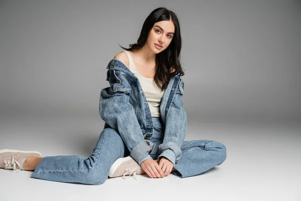 Full length of young alluring woman with long brunette hair and flawless natural makeup posing in stylish blue jeans and denim jacket while sitting on grey background — Stock Photo
