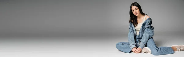 Полная длина очаровательной модели с длинными волосами брюнетки и безупречным естественным макияжем позируя в стильных синих джинсах и джинсовой куртке, сидя на сером фоне, баннер — стоковое фото