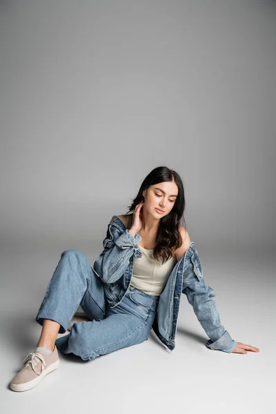 Volle Länge der verträumten jungen Frau mit langen brünetten Haaren und makellosem natürlichen Make-up posiert in stilvollen blauen Jeans und Jeansjacke, während sie auf grauem Hintergrund sitzt — Stockfoto