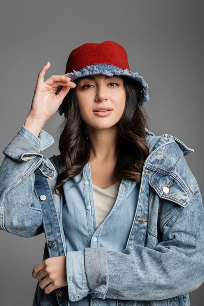 Portrait of young alluring woman with flawless natural makeup posing in panama hat and denim jacket and looking at camera on grey background — Stock Photo