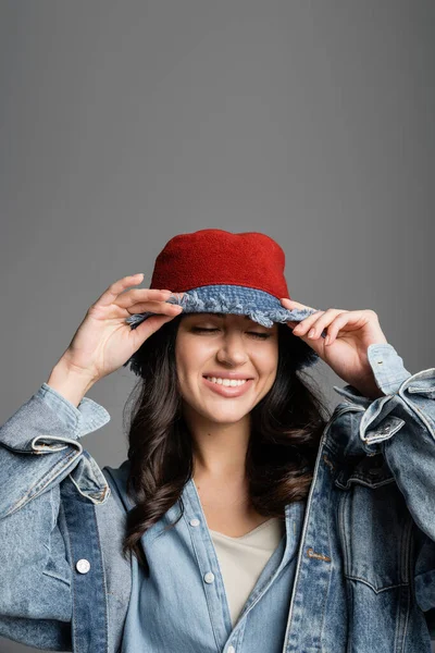 Retrato de mujer encantadora joven con maquillaje natural impecable posando en sombrero de panama y chaqueta de mezclilla mientras sonríe sobre fondo gris - foto de stock
