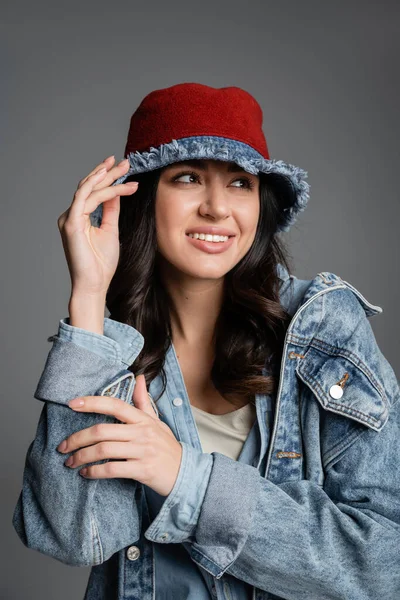 Портрет усміхненої молодої жінки з бездоганним натуральним макіяжем позує в панамському капелюсі і джинсовій куртці, дивлячись на сірий фон — стокове фото