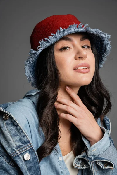 Retrato de jovem mulher encantadora com maquiagem natural impecável posando em chapéu panamá e jaqueta de ganga e olhando para a câmera em fundo cinza — Fotografia de Stock