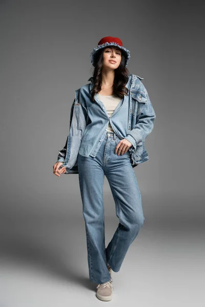 Полная длина молодая очаровательная женщина с безупречным естественным макияжем позируя в джинсовой шляпе Панама, синие джинсы и куртка стоя и глядя на камеру на сером фоне — стоковое фото