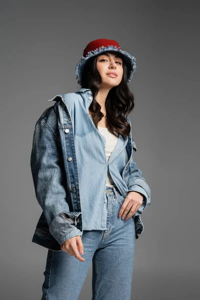 Молодая очаровательная женщина с безупречным естественным макияжем позирует в стильной панамской шляпе и джинсовой куртке, глядя в камеру и стоя на сером фоне — стоковое фото