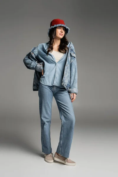 Полная длина молодой очаровательной женщины с безупречным естественным макияжем позируя в джинсовой шляпе, синих джинсах и куртке, стоя с рукой на бедре на сером фоне — стоковое фото
