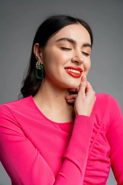 Portrait of happy young woman with closed eyes, shiny brunette hair, trendy earrings, red lips and stylish magenta dress smiling while posing isolated on grey background — Stock Photo