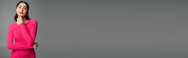 Incantevole giovane donna con capelli castani lucidi, orecchini alla moda, labbra rosse ed elegante abito magenta guardando la fotocamera e posando su sfondo grigio, banner — Foto stock