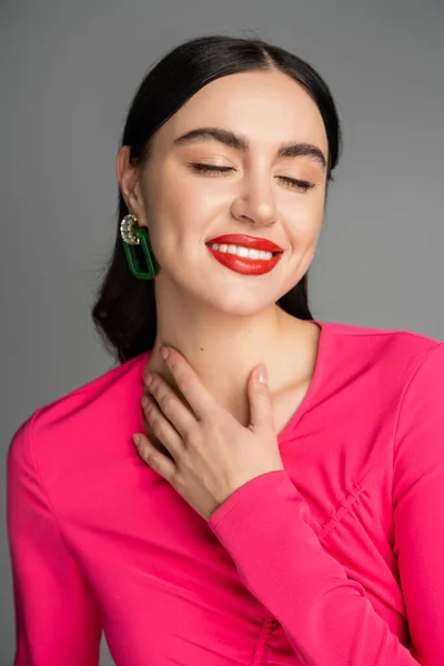 Portrait of pleased woman with shiny brunette hair, trendy earrings, red lips and stylish magenta dress smiling with closed eyes isolated on grey background — Stock Photo