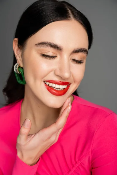 Портрет счастливой молодой женщины с блестящими брюнетками, модными сережками и красными губами, улыбающимися с закрытыми глазами, позируя на сером фоне — стоковое фото