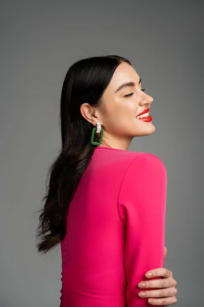 Счастливая молодая женщина с брюнетками, модные серьги, красные губы и стильное пурпурное платье улыбается с закрытыми глазами и позирует на сером фоне — стоковое фото