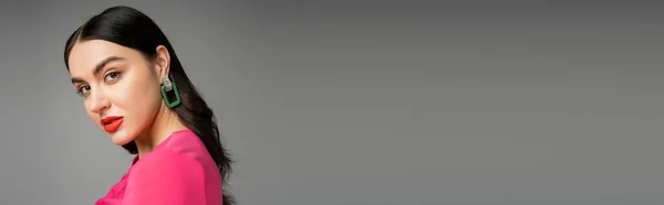 Encantadora jovem com cabelo morena, brincos na moda, lábios vermelhos e vestido magenta elegante olhando para a câmera e posando em fundo cinza, banner — Fotografia de Stock