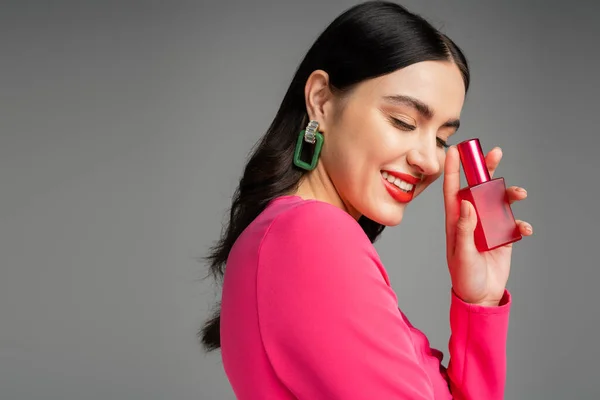 Chic young woman with brunette hair, trendy earrings, red lips and stylish magenta dress holding bottle of luxurious perfume and smiling on grey background — Stock Photo