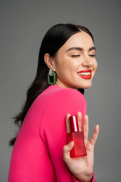Pleased woman with brunette hair, trendy earrings, red lips and stylish magenta dress holding bottle of luxurious perfume and smiling on grey background — Stock Photo