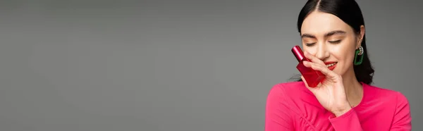 Donna glamour con capelli castani, orecchini alla moda, labbra rosse ed elegante abito magenta bottiglia di profumo di lusso e sorridente su sfondo grigio, banner — Foto stock