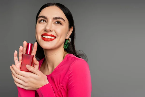 Mujer glamorosa con el pelo moreno labios rojos y vestido magenta de moda sosteniendo botella de perfume de lujo y mirando hacia otro lado, sonriendo sobre fondo gris - foto de stock