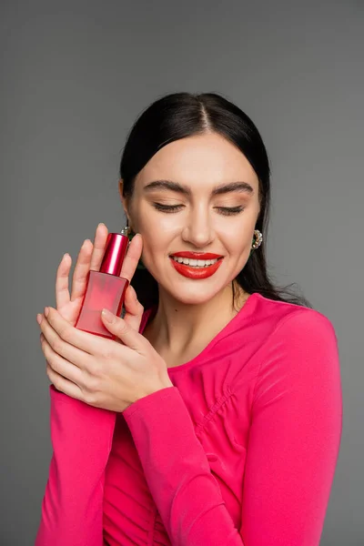 Retrato de una hermosa joven con cabello moreno y labios rojos posando en vestido magenta mientras sostiene la botella de perfume de lujo y sonriendo aislado sobre fondo gris - foto de stock