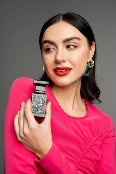 Portrait of alluring young woman with shiny brunette hair, red lips and magenta dress holding bottle of perfume and looking away on grey background — Stock Photo