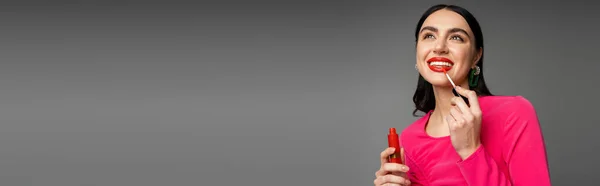 Splendida giovane donna con capelli castani e orecchini alla moda in posa in abito da festa magenta mentre si applica lucidalabbra rosso e sorridente isolato su sfondo grigio, banner — Foto stock