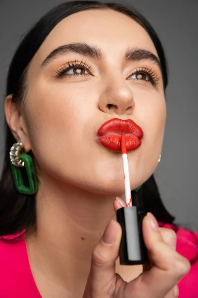 Vista de cerca de la mujer joven con pelo moreno brillante y pendientes de moda pucheros labios mientras se aplica brillo labial rojo y posando aislado sobre fondo gris - foto de stock
