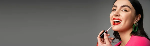 Donna seducente con orecchini alla moda e capelli castani applicando lucidalabbra rosso e sorridente mentre guarda la fotocamera e posa isolata su sfondo grigio, banner — Foto stock