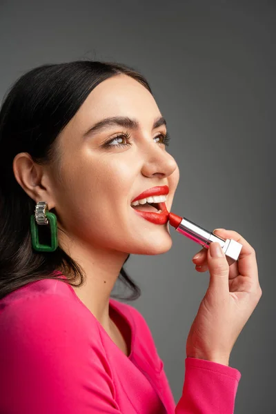 Alluring woman with trendy earrings and shiny brunette hair applying red lipstick and smiling while looking away and and preparing for party isolated on grey background — Stock Photo