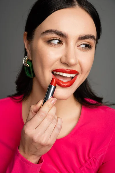 Charming woman with trendy earrings and shiny brunette hair applying red lipstick and smiling while looking away and and preparing for party isolated on grey background — Stock Photo