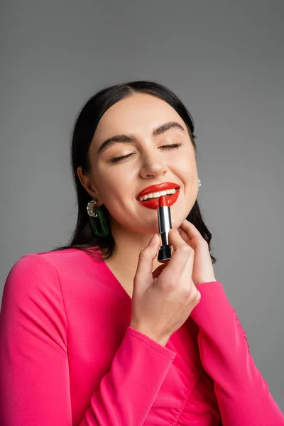 Alluring woman with trendy earrings and shiny brunette hair applying red lipstick and smiling with closed eyes while and preparing for party isolated on grey background — Stock Photo