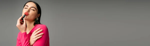 Divertente donna con orecchini alla moda e capelli lucidi bruna applicare rossetto rosso e labbra pouting mentre posa isolata su sfondo grigio, banner — Foto stock