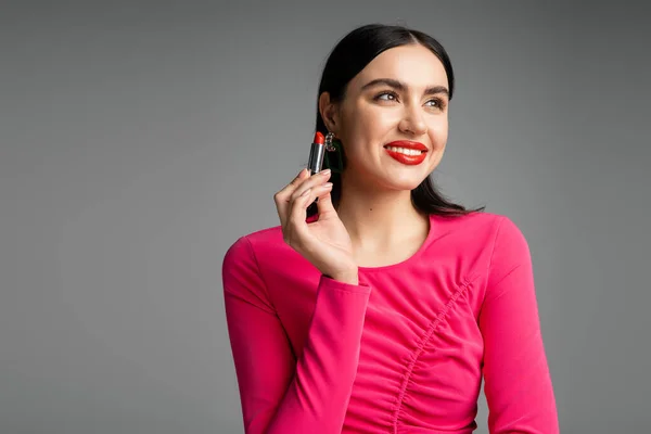 Happy woman with trendy earrings and shiny brunette hair holding red lipstick and smiling while looking away and posing isolated on grey background — Stock Photo