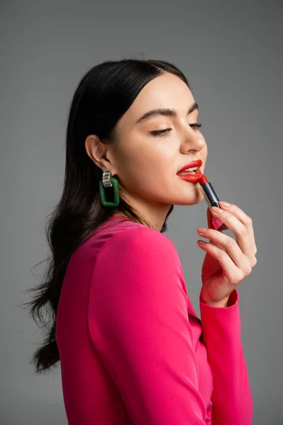 Young and charming woman with trendy earrings and shiny brunette hair posing while applying red lipstick and looking away isolated on grey background — Stock Photo