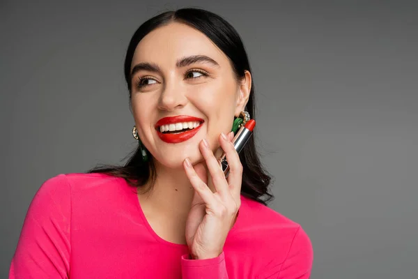 Portrait of elegant woman with trendy earrings and shiny brunette hair holding red lipstick between fingers and smiling while posing on grey background — Stock Photo