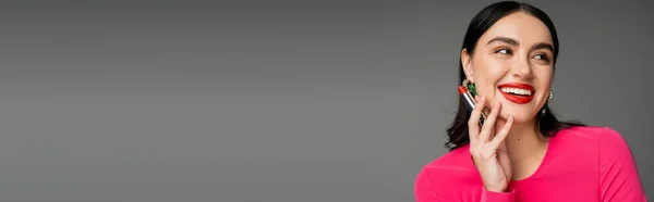 Porträt einer schönen Frau mit trendigen Ohrringen und brünetten Haaren, die roten Lippenstift zwischen den Fingern hält und lächelt, während sie auf grauem Hintergrund posiert, Banner — Stockfoto